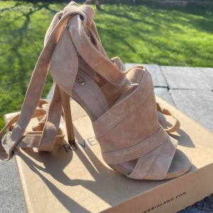 Beiga mockasandaler med klack River Island - Snygga sandaler i beige mocka från River Island med hög klack och öppen tå. Skorna har breda remmar som korsar över foten och långa knytband som snurras runt vristen för en trendig look. Perfekta för att lyfta din outfit till nästa nivå.