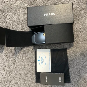 Svarta solglasögon från Prada - Snygga svarta solglasögon från Prada med breda, geometriska skalmar och tydlig logga på sidan. Glasögonen har en modern, kantig form och kommer med originalfodral, putsduk och certifikat. Perfekta för dig som vill sticka ut med en lyxig accessoar.