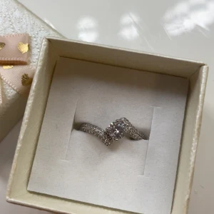 Silverring med glittrande stenar - Säljer en snygg rostfritt silverfärgad ring med en rund, gnistrande sten i mitten och mindre stenar längs bandet. Ringen har en elegant, lätt böjd form som ger en unik look. 