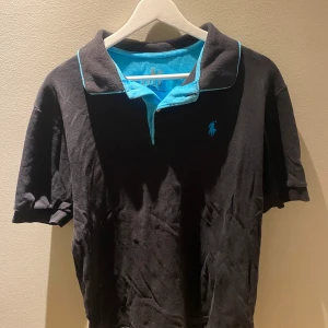 Svart pikétröja från Polo - Säljer en svart pikétröja från Polo med turkosa detaljer. Tröjan har korta ärmar och en klassisk krage med turkos insida och logotyp. Perfekt för en stilren look.