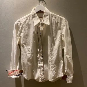 Vit skjorta från Burberry Brit - Snygg vit skjorta från Burberry Brit med klassisk krage och knappar framtill. Skjortan har långa ärmar och en diskret broderad logga på bröstet. Insidan av manschetterna har ett stilrent rutigt mönster. Storlek M men passar S