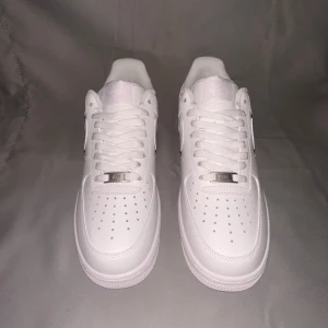 Vita Nike Air Force sneakers - Snygga och klassiska vita Nike Air Force sneakers med snörning. Perfekta för en stilren look. Skorna har en ikonisk design med perforerade detaljer på tån och en robust sula för extra komfort.