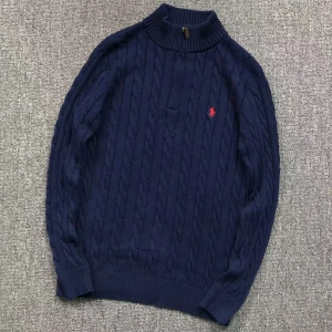 Mörkblå stickad tröja från Polo Ralph Lauren - Säljer en snygg mörkblå stickad tröja från Polo Ralph Lauren med kabelstickat mönster. Tröjan har en broderad röd logotyp på bröstet och ribbstickad krage samt ärmslut. Perfekt för kyligare dagar!