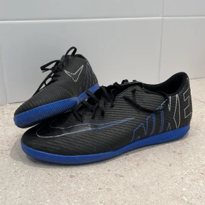 Svarta och blå Nike fotbollsskor - Ett par Nike Mercurial vapor 15 futsal skor, skick 9/10 använda en sesong pågrund av jag växte ur dom värligt snabbt. Perfekta för den som vill ha stil och funktion på planen. Skorna tvättas innan köp. Skriv för mer bilder eller information.