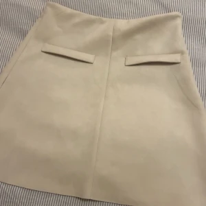 Beige kjol från Zara - Snygg beige kjol från Zara i en stilren design. Kjolen har en dragkedja på sidan och dekorativa fickdetaljer framtill. Kjolen har två små hål i sömnen på ena sidan men som lätt går att sy ihop! 💗