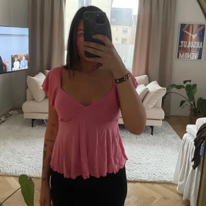 Rosa topp med knytning - Säljer en söt rosa topp med v-ringning och volangdetaljer. Toppen har korta ärmar och en fin knytning i ryggen som ger en extra touch. Perfekt för en avslappnad stil.
