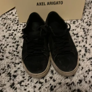 Svarta sneakers från Axel Arigato - Snygga svarta sneakers från Axel Arigato med klassisk design och snörning. Perfekta för en stilren look. Skorna har en vit sula som ger en fin kontrast till det svarta.