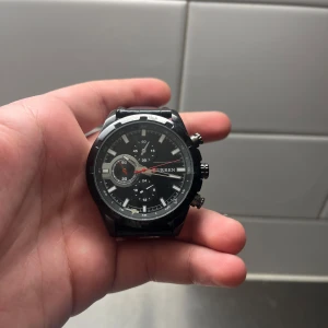 Svart herrklocka från Curren - Snygg svart herrklocka från Curren med en stilren urtavla och flera små urtavlor för extra funktioner. Klockan har en svart läderrem och silverdetaljer som ger en elegant look. Perfekt för den som vill ha en stilren accessoar.