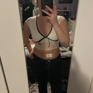 Vit spetstopp - Säljer en snygg vit spetstopp med svarta kanter och knytning framtill. Toppen är kortärmad och har en djup v-ringning. Perfekt för att ge en elegant touch till din outfit. Aldrig använd så är i nyskick