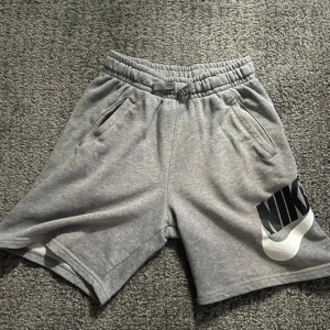 Grå shorts från Nike - Säljer ett par grå Nike shorts med elastisk midja och fickor. De har en stor Nike-logga i svart och vitt på ena benet. Perfekta för en avslappnad stil.