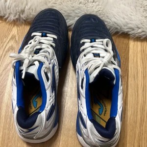 Blå och vita sneakers från Joma - Snygga sneakers från Joma i blått och vitt med snörning. Skorna har en sportig design med blå detaljer och en robust sula. Perfekta för en aktiv livsstil. Priset kan diskuteras, har inte använt den 