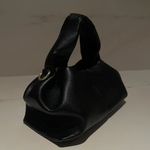 Svart handväska med axelrem - Snygg svart handväska i skinn imitation med en avtagbar axelrem. Väskan har en stilren design med guld detaljer. Perfekt för att bära både i handen och över axeln.