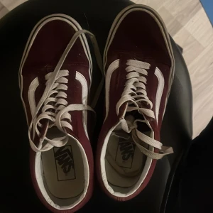 Vinröda sneakers från Vans - Säljer ett par klassiska vinröda Vans sneakers med vita detaljer och snörning. Skorna har den ikoniska vita sidoremsan och en vit sula med 'Off the Wall' logga på hälen. Perfekta för en avslappnad stil.