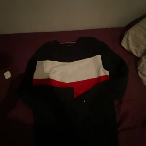 Svart tröja från Tommy Hilfiger - Säljer en svart tröja från Tommy Hilfiger med en bred vit och röd rand över bröstet. Tröjan har långa ärmar och en rund halsringning. Perfekt för en stilren och avslappnad look.