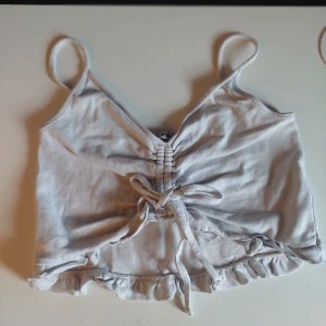 Vit linnetopp med volangdetaljer - Säljer en söt vit linnetopp med smala axelband och volangdetaljer i nederkant. Toppen har en knytning framtill som ger en fin detalj och en lätt croppad passform. Perfekt för varma sommardagar!