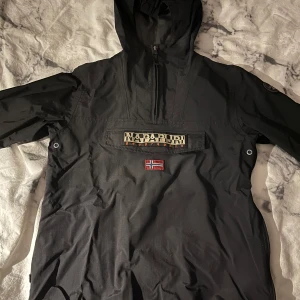 Svart vindjacka från Napapijri - Säljer en stilren svart halfzip vindjacka från Napapijri med huva. Jackan är tunn men passar perfekt för blåsiga och regniga dagar. 