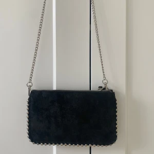 Svart axelväska med kedja - Snygg svart axelväska i mocka med silverfärgad kedja och dragkedja. Väskan har dekorativa nitar längs kanterna som ger en cool look. Perfekt för att lyfta din outfit!