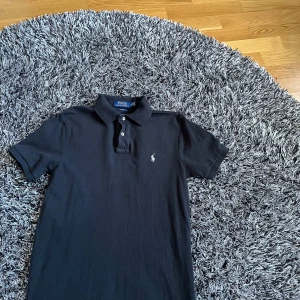 Svart pikétröja från Ralph Lauren - Säljer en klassisk svart pikétröja från Ralph Lauren i storlek M. Tröjan har en broderad logga på bröstet och knappar vid kragen. Perfekt för en stilren look. Skick: 9/10 Nypris runt 1500kr