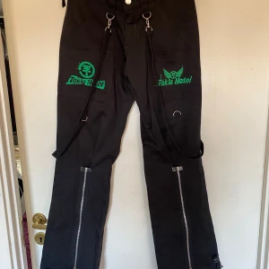 Tokio hotel byxa storlek S.  - Unika svarta byxor med gröna broderade Tokio Hotel-loggor. Byxorna har dragkedjor längs benen och flera dekorativa remmar och ringar. Perfekta för fans av bandet som vill ha en edgy stil. En liten ficka med dragkedja på ena benet. Material i bomull. Endast provade. Stretchiga. Midjevidd 76 cm. Innerbenslängd ca 81 cm.