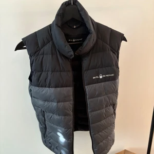 Svart quiltad dunväst från Sail Racing - Snygg svart quiltad dunväst från Sail Racing med dragkedja framtill. Västen har en hög krage och logotyp på bröstet och ryggen. Perfekt för kyliga dagar.