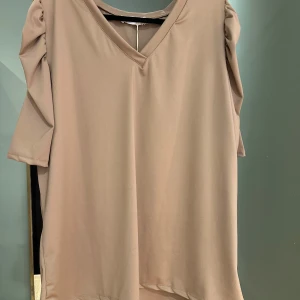 Beige topp från Gemini - Säljer en stilren beige topp från Gemini med v-ringning och korta ärmar. Tillverkad i en mjuk och stretchig blandning av polyester och elastan. Perfekt för en avslappnad look.
