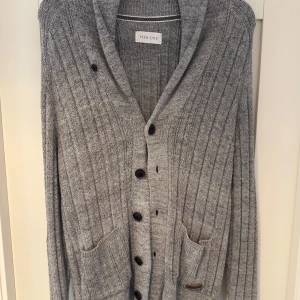 Grå ribbad kofta från Pier One - Säljer en stilren grå ribbad kofta från Pier One med knappar framtill och praktiska fickor. Koftan har långa ärmar och bruna armbågslappar som ger en snygg kontrast. 