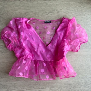 Rosa prickig blus - Säljer en söt rosa blus med prickigt mönster och v-ringning. Blusen har korta puffärmar och volangdetaljer vid midjan. Perfekt för en färgglad look!