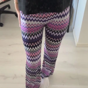 Färgglada mönstrade byxor Alice by shoes alice  - Säljer ett par färgglada byxor med zigzag-mönster i lila, rosa, svart och vitt. Byxorna har en bekväm passform och är perfekta för att liva upp garderoben. De är tillverkade i ett mjukt och stretchigt material. S/M