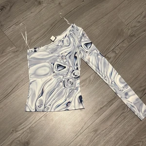 Mönstrad oneshoulder-topp - Snygg oneshoulder-topp med abstrakt mönster i blått och vitt. Toppen har en lång ärm och en unik design som verkligen sticker ut. Perfekt för att ge din outfit en cool touch. 