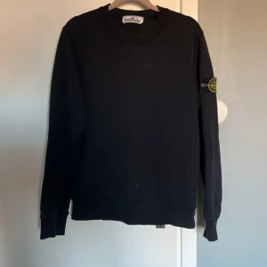 Svart tröja från Stone Island - Säljer en stilren svart tröja från Stone Island med deras ikoniska märke på ärmen. Tröjan har långa ärmar och en klassisk rund halsringning. Perfekt för en casual look.