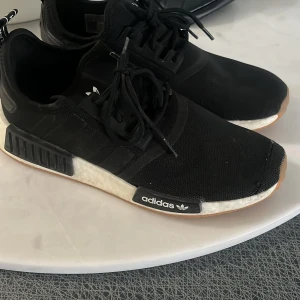 Svarta Adidas NMD sneakers - Säljer ett par svarta Adidas NMD sneakers med vit sula och klassisk tre-randig design. Skorna har snörning och är tillverkade i syntetmaterial. Perfekta för en stilren och bekväm look.