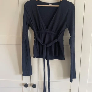 Mörkblå ribbad omlottopp - Jag säljer den populära Nelly tröjan. Det är en mörkblå ribbad omlottopp med långa ärmar och knytband i midjan. Har inte använts någon gång men kommer inte till användning 