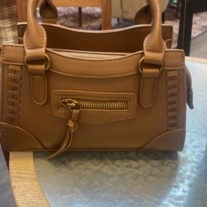 Brun handväska från Gina Tricot - Säljer en snygg brun handväska från Gina Tricot. Väskan har en praktisk dragkedja framtill och stiliga detaljer. Perfekt för att bära dina nödvändigheter med stil.❤️