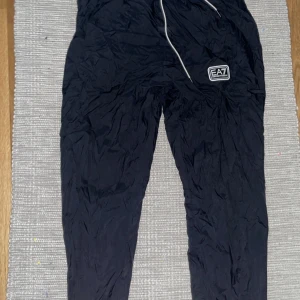 Intressekoll 03 - Snygga svarta träningsbyxor från EA7 med elastisk midja och snörning. Perfekta för träning eller en avslappnad dag. Byxorna har en logotypdetalj på benet och är tillverkade i ett lätt syntetmaterial.