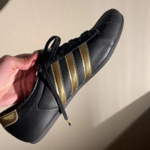 Adidas skor  - Snygga svarta sneakers från Adidas med guldiga detaljer och de klassiska tre ränderna på sidan. Skorna har snörning och en unik mönstrad insida i svart och guld. Perfekta för en stilren look. Passar både tjej och kille då det beror på din storlek😁Helt oanvända med prislapp på 