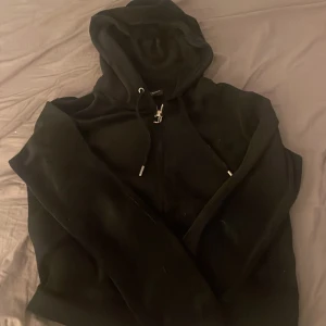 Svart Juicy Couture hoodie i velour - Svart hoodie från Juicy Couture i mjuk velour med dragkedja och snörning i huvan. Klassisk JC-logga broderad på bröstet och snygga metalldetaljer på snörena. Perfekt för dig som gillar en chill och trendig look.