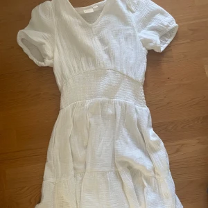 Vit kortklänning från Gina Tricot - Supersöt vit kortklänning från Gina Tricot med v-ringning, puffiga korta ärmar och smockad midja. Tillverkad i mjuk bomull med lätt struktur och volangig kjol. Perfekt för varma dagar och en fräsch look.