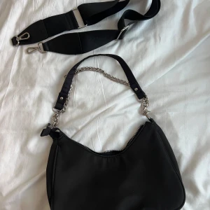 Svart axelväska med kedjedetalj - Säljer en svart axelväska med cool kedjedetalj på axelremmen och dragkedja upptill. Väskan har en mjuk form och extra bred, justerbar rem som kan bytas ut. Perfekt storlek för det viktigaste och snygg att bära crossbody eller på axeln.