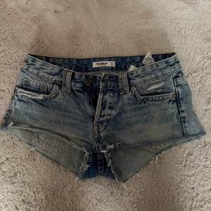 Blå lågmidjade jeansshorts  - Snygga ljusblå jeansshorts från Pull&Bear med råa kanter och slitna detaljer. Klassisk femficksmodell med bälteshällor och knappgylf. Perfekta för varma dagar och en avslappnad stil.