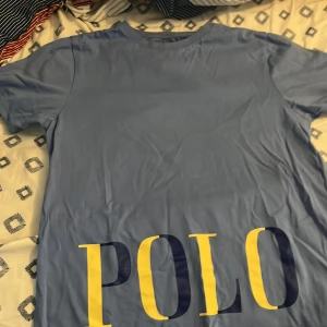 Blå t-shirt från Polo Ralph Lauren - Snygg blå t-shirt från Polo Ralph Lauren med stor POLO-logga i gult och mörkblått framtill. Klassisk rund hals och korta ärmar. Skön och mjuk bomull som passar perfekt till jeans eller shorts. Det står 14- 16 men skulle säga att den passar S perfekt 