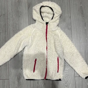 Vit teddyjacka med huva från XTTL för barn - Mysig vit teddyjacka från XTTL i storlek 120. Jackan har huva, långa ärmar och två fickor med rosa dragkedjor. Perfekt för kyliga dagar när du vill vara både varm och stilren. Den fluffiga looken ger en trendig vibe och jackan stängs med rosa dragkedja framtill.