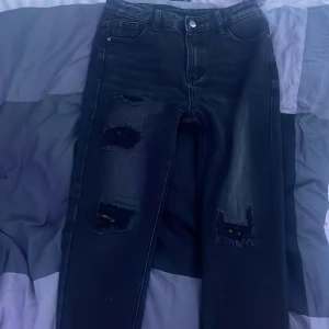 Svarta ripped jeans med raka ben - Säljer ett par svarta jeans med snygga slitningar på båda benen. Jeansen har raka ben, klassiska fem fickor och är tillverkade i en mix av bomull och polyester. Perfekta för dig som gillar en lite tuffare stil.