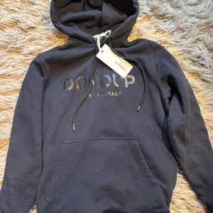 Dondup hoodie - Svart hoodie från Dondup med snyggt logotyptryck på bröstet. Klassisk modell med huva, snörning och stor känguruficka framtill. Tillverkad i mjuk bomull som känns skön mot huden. Storlek M. Ny skick 10/10 skick. Bara att skriva vid minsta fundering