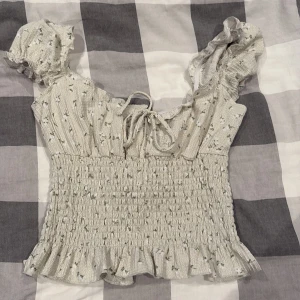 Blommig blus med volang från Gina Tricot - Supersöt ljus blus från Gina Tricot med små blommor i grönt och vitt. Toppen har volangkant nertill, smockad midja, puffiga korta ärmar och knytning fram. Perfekt för en romantisk look och passar till både jeans och kjol.