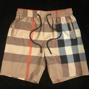Burberry rutiga shorts med snörning - Snygga shorts från Burberry med klassiskt rutigt mönster i beige, svart, vitt och rött. De har elastisk midja med svart snörning och sidofickor. Perfekta för sommaren och ger en lyxig vibe till din outfit.