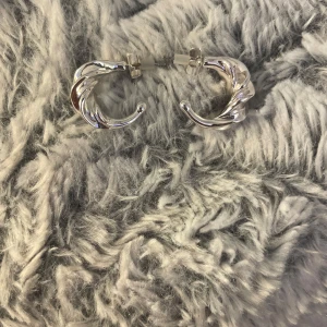 Silvriga chunky örhängen i tvinnad design - Säljer ett par chunky örhängen i silverfärg med tvinnad, snirklig form. De är halvcirkelformade och har stift med pluppar. Perfekta för dig som gillar statement-smycken med modern känsla.