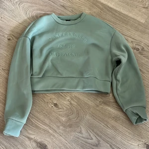 Grön croppad sweatshirt San Francisco - Snygg ljusgrön croppad sweatshirt med tryck 'San Francisco California' framtill. Tröjan har rund hals, långa ärmar och bred mudd nertill. Perfekt för en chill och avslappnad stil.