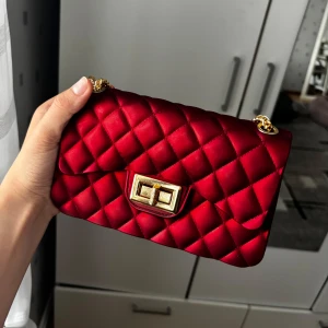 Röd quiltad axelväska med guldkedja - Snygg röd axelväska med quiltat mönster och guldfärgad kedja. Väskan har ett vridlås i guld och ett rymligt fack inuti. Perfekt för att lyfta din outfit med en pop av färg och lyxig känsla.