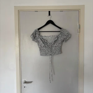 Grönvit croppad blus med leopardmönster - Supersöt croppad blus i kall grön och vitt med leopardmönster. Puffiga korta ärmar, v-ringning och smockad midja. Snygg snörning framtill och volangdetaljer vid axlarna. Perfekt för dig som vill sticka ut lite extra!