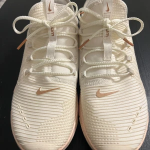 Nike Zoom träningssneakers cremevit/rosa - Nike Zoom sneakers i cremevit med detaljer i ljusrosa och metallisk rosa. Snyggt ribbad ovandel, snörning och Nike-logga på framsidan. Sulan är ljusrosa med grått mönster undertill. Perfekta för träning och gym, riktigt fräsch och modern look.